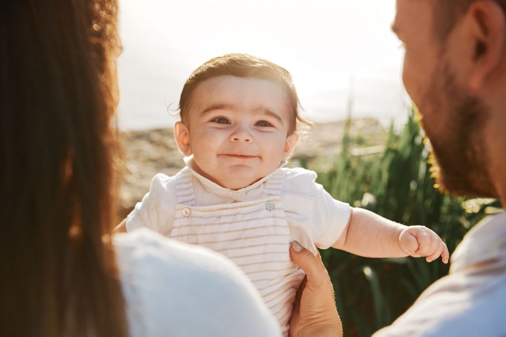 Adorable smiling baby held by parents in warm golden sunlight outdoors.