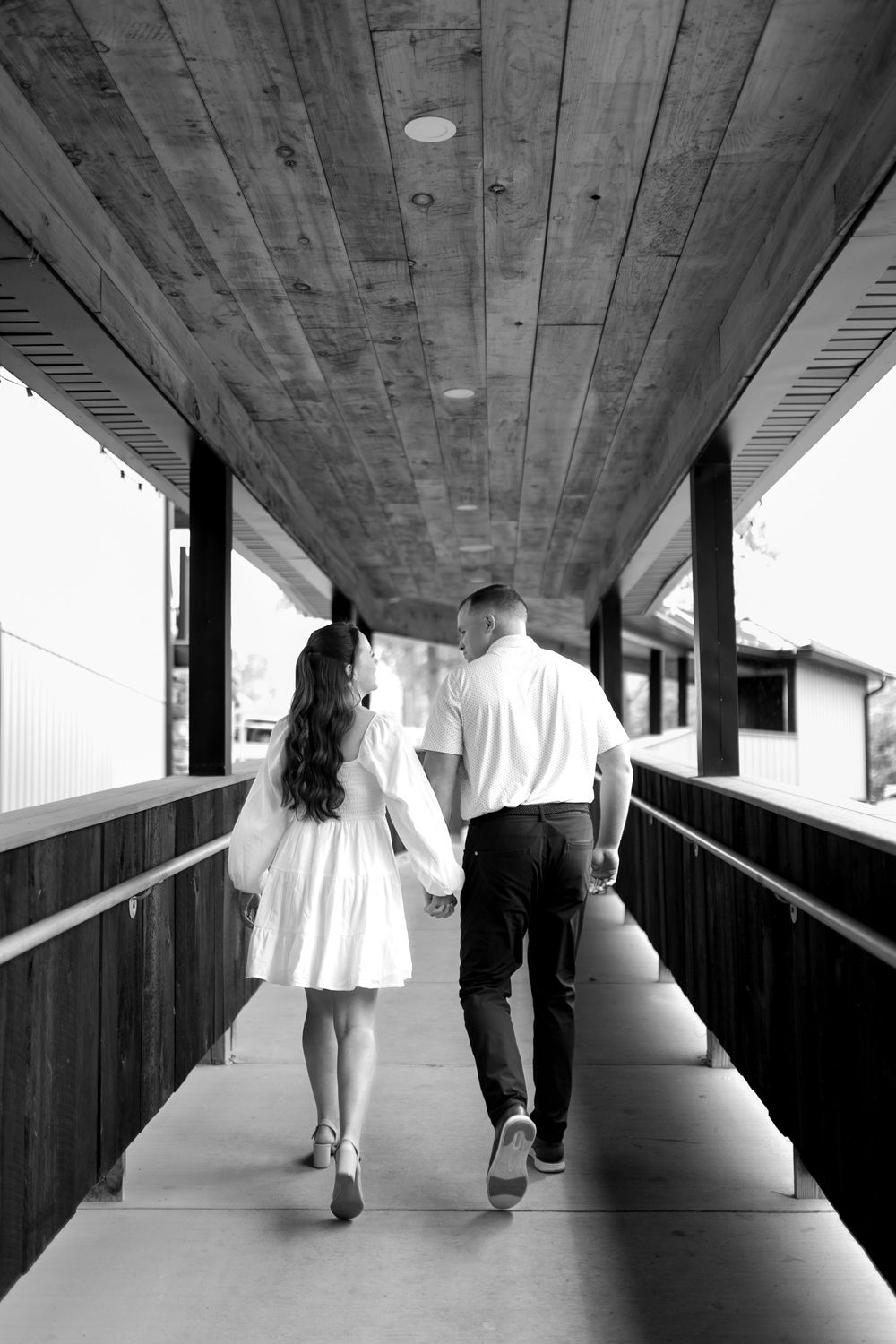 A couple walking and holding hands on a covered wooden walkway, captured in black and white.