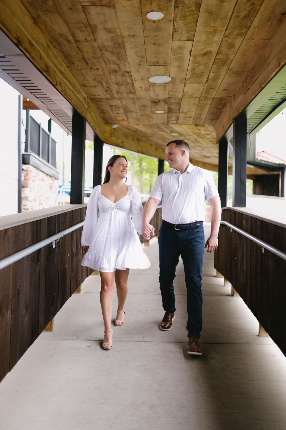 A couple wearing white and dark clothing walks together in a covered wooden walkway with natural lighting.
