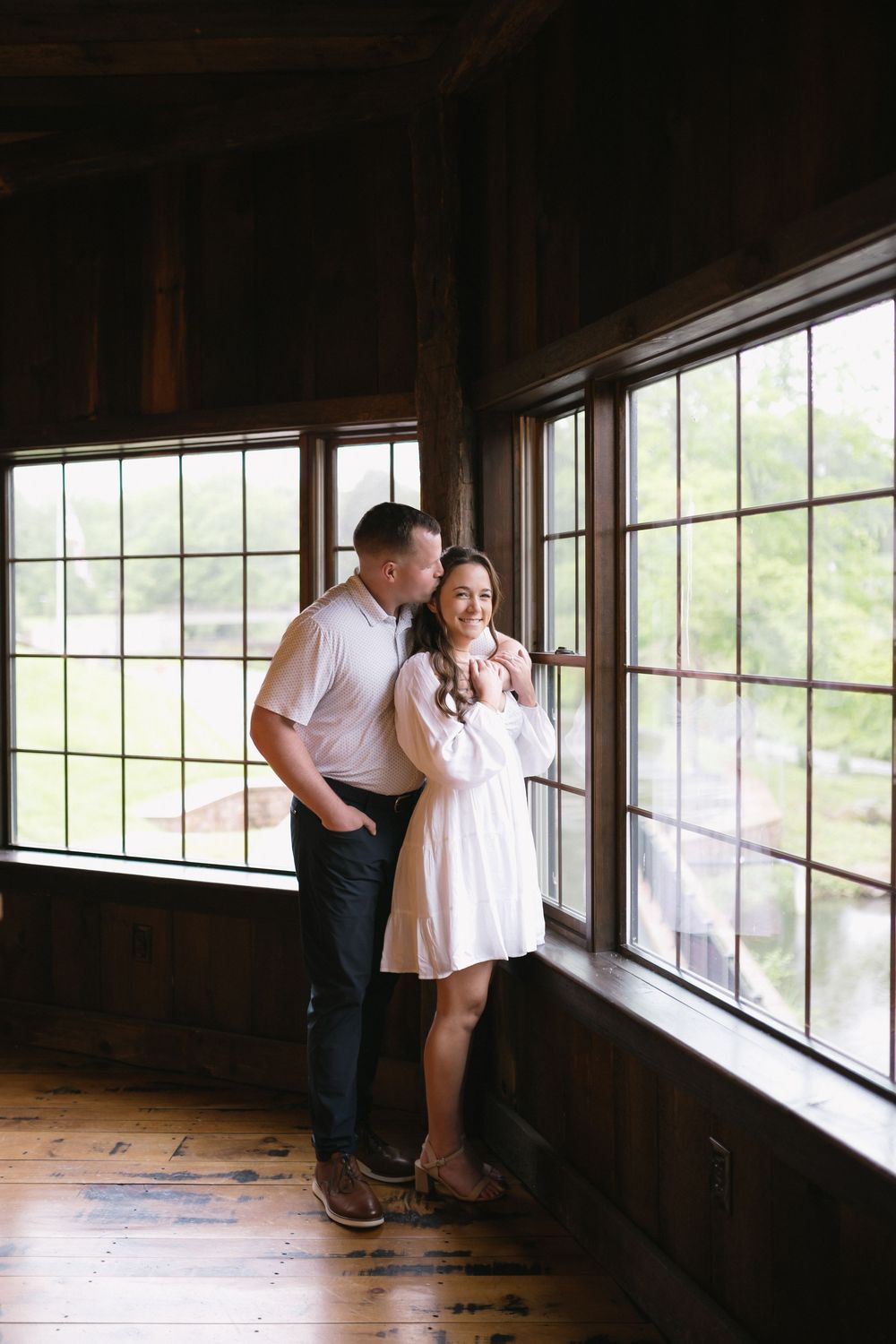 Couple in white clothing stands together by large windows in a rustic wooden interior.