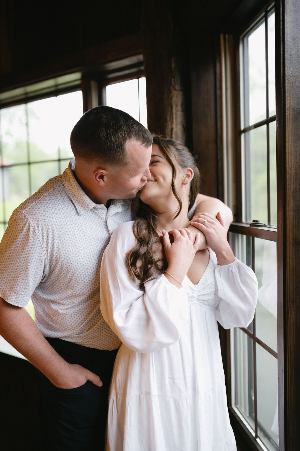 A romantic moment shared by a couple in white and black attire standing by large windows in natural light.