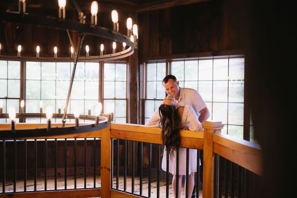 An individual leans against a wooden railing inside a rustic lodge-style room with large windows and chandelier lighting.