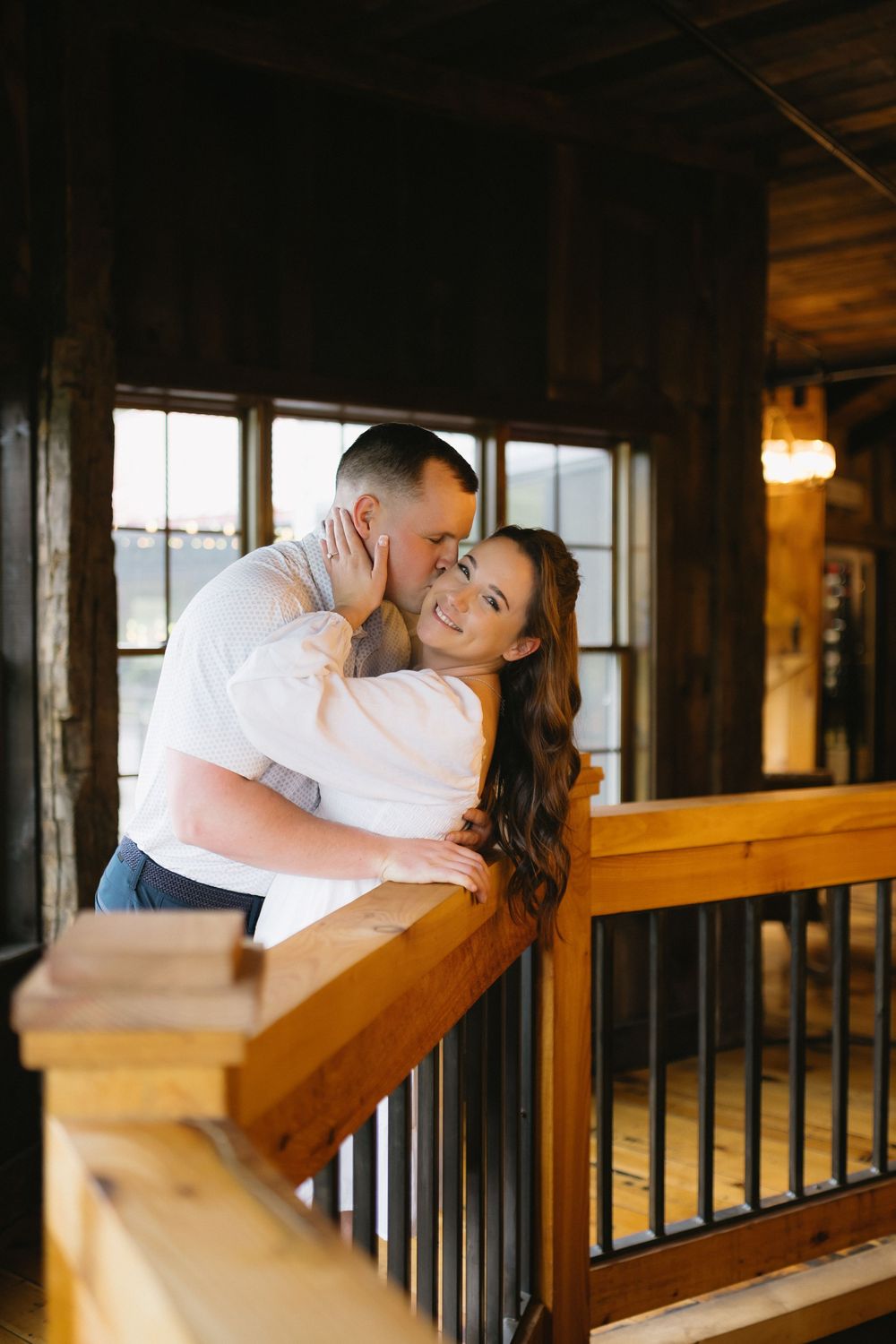 A romantic couple shares an intimate moment on a rustic wooden balcony with warm lighting and large windows.