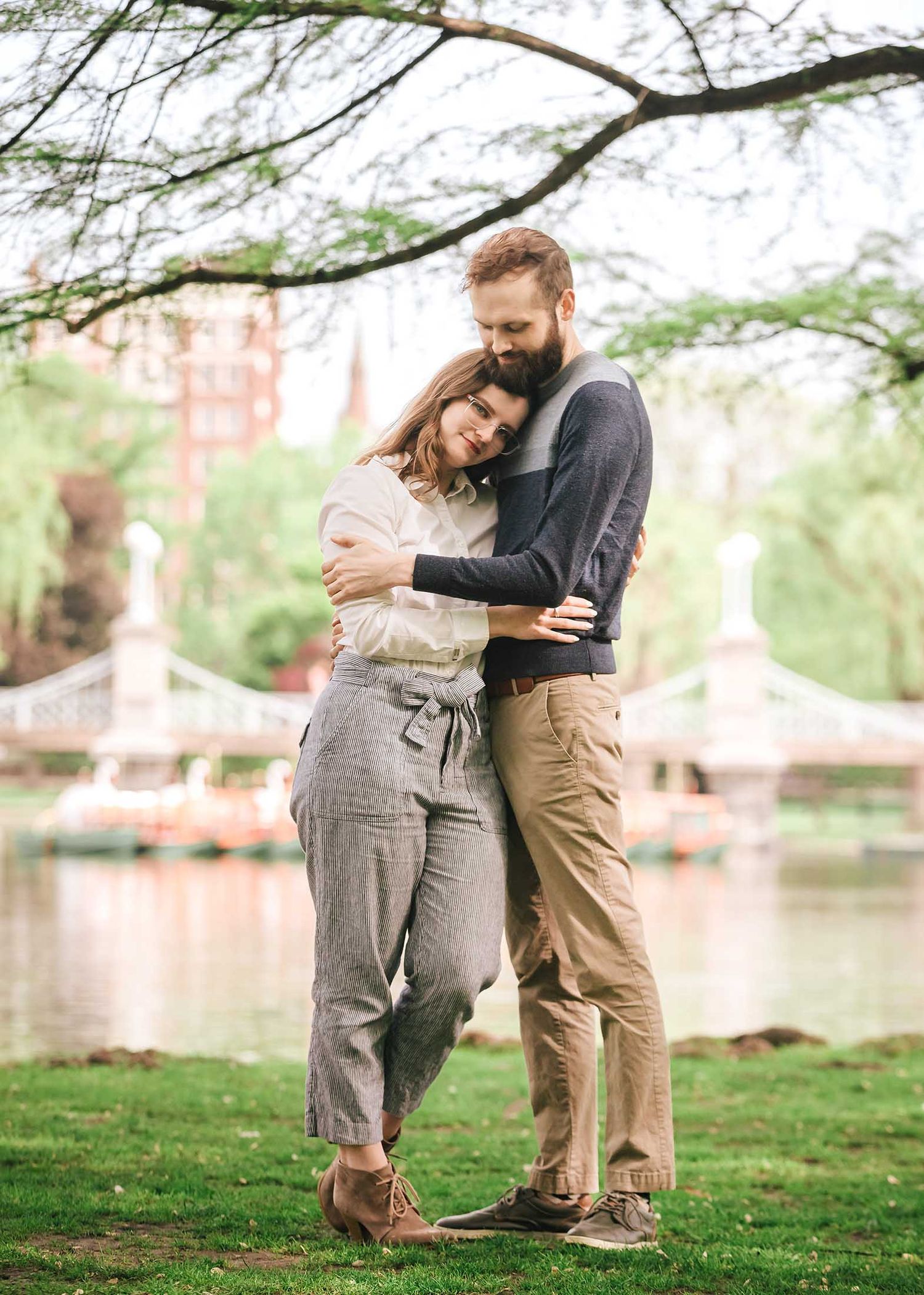 Tender moment of engaged couple cuddling under sweeping branches with Boston Public Garden Foot Bridge in the background