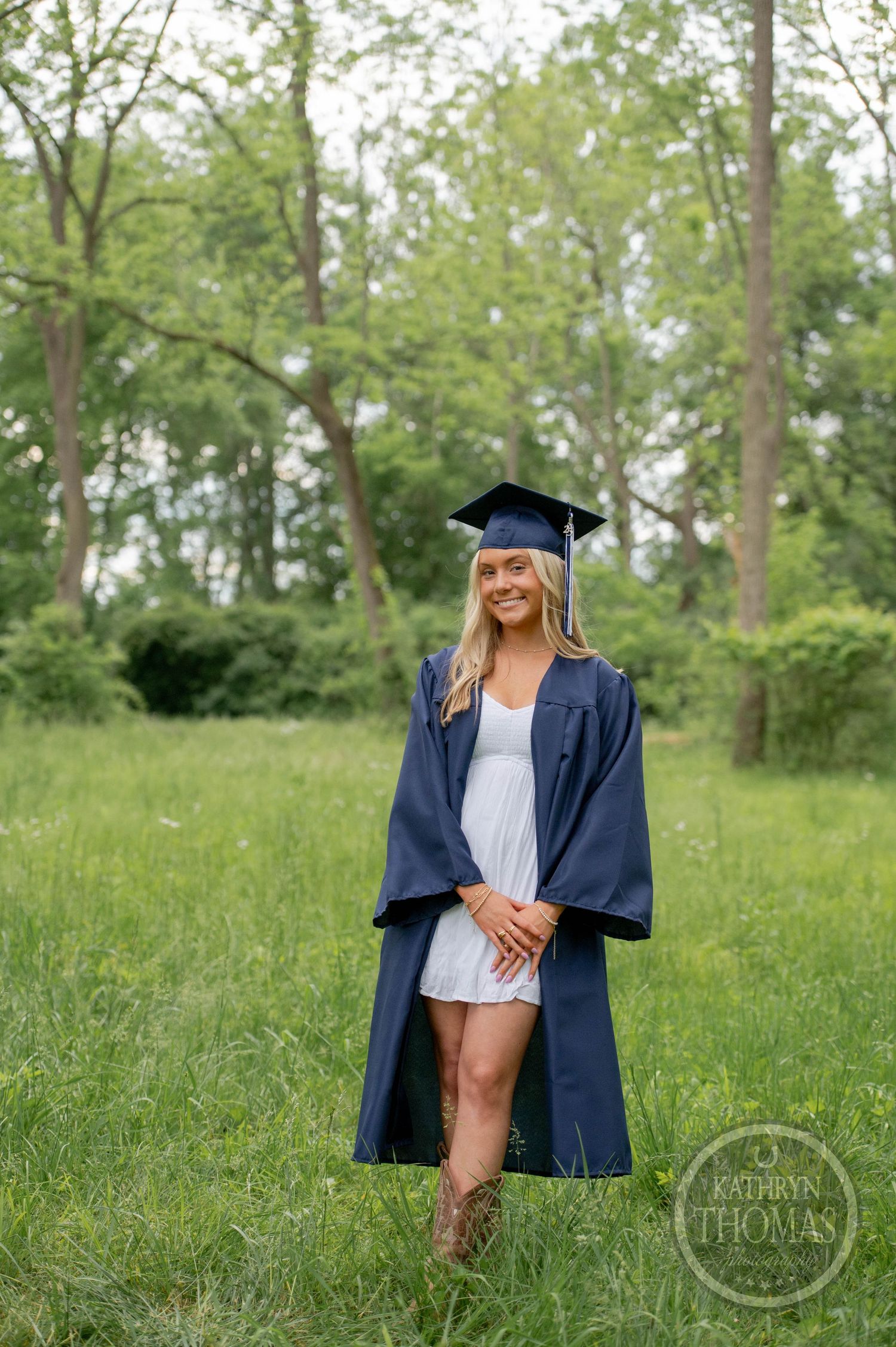 Graduate in navy blue cap and gown stands in green field with trees in background during outdoor photo session.