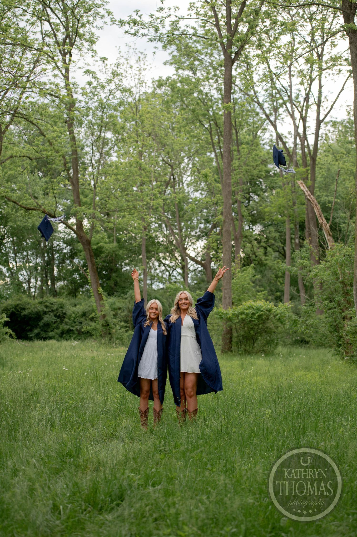 Two students in graduation attire jump joyfully in a lush green field surrounded by trees.