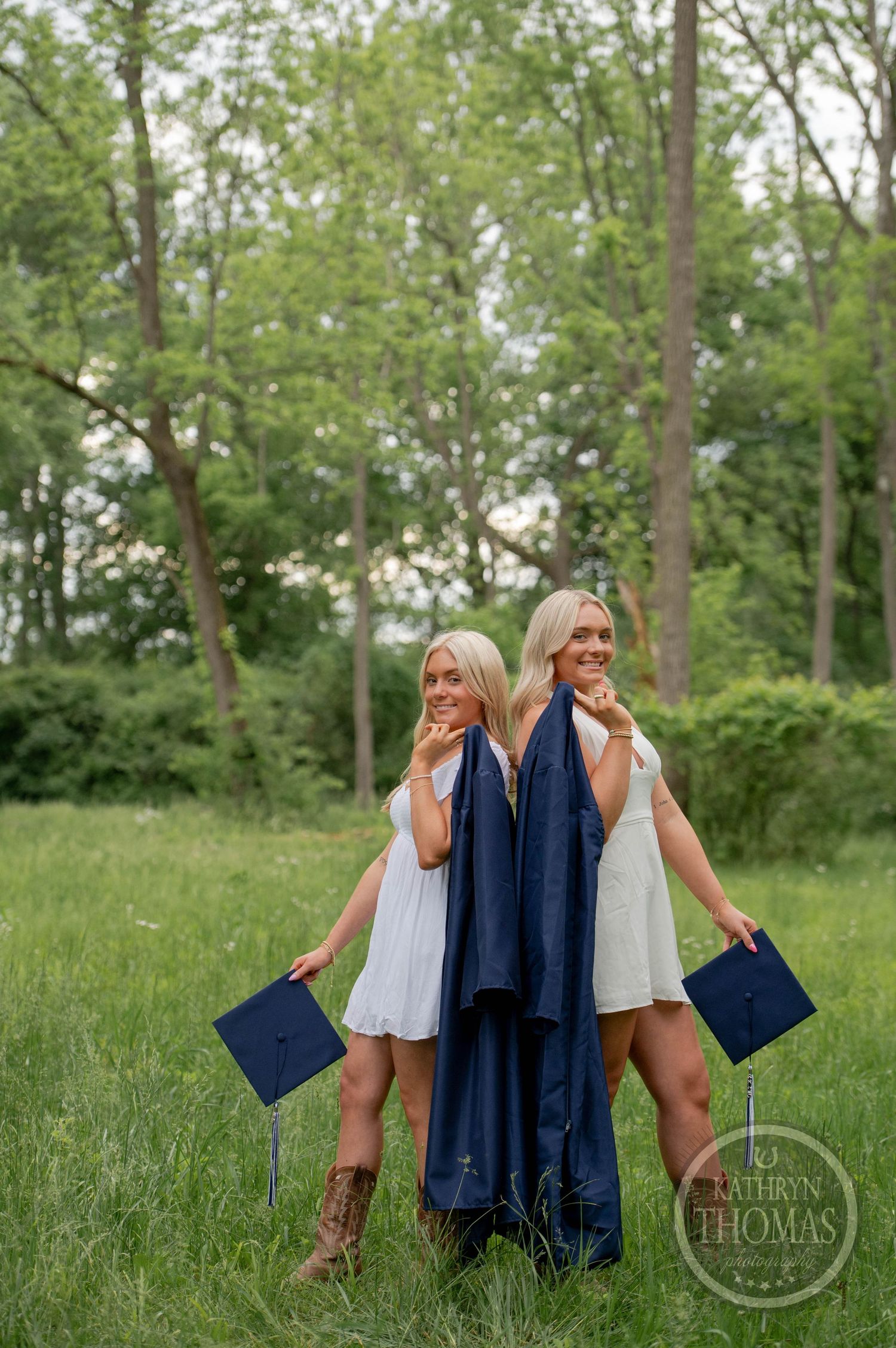 Two graduates in white dresses pose back-to-back with navy gowns and diplomas in a grassy outdoor setting.