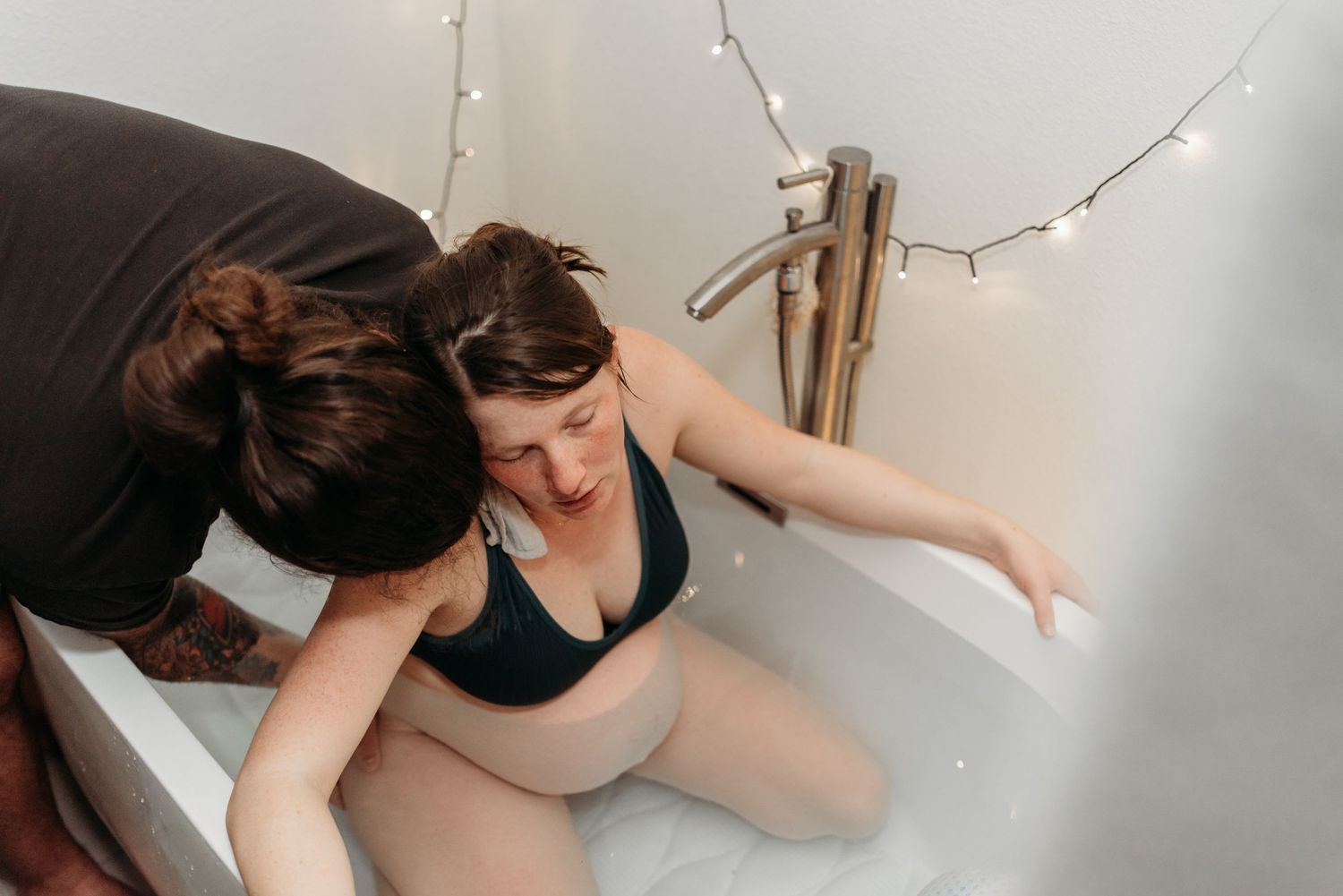Two friends sharing an emotional moment together while sitting in a bathtub with string lights in the background.