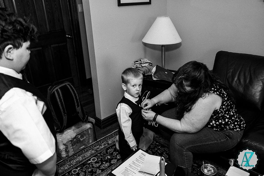 A candid black and white moment captured during a formal preparation scene showing adjustments being made to a young vest.