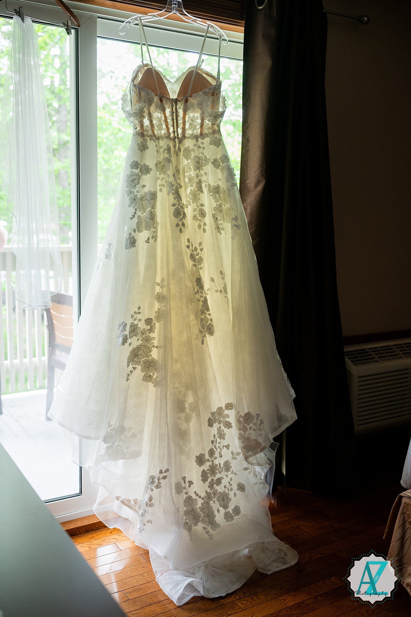 Several delicate white wedding dresses with intricate lace details and floral embellishments hang in a sunlit room.