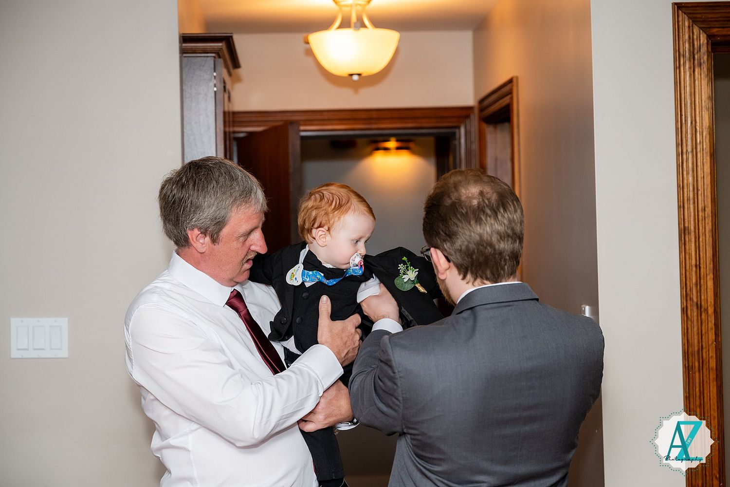 Two adults in formal attire helping dress a small child in a hallway under warm lighting.