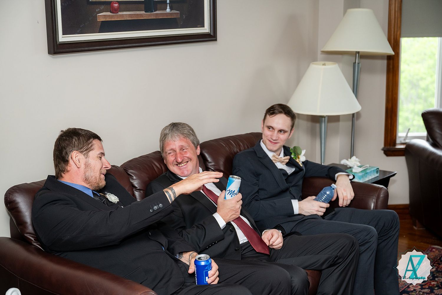 Three people laughing while relaxing together on a brown leather couch in a living room.