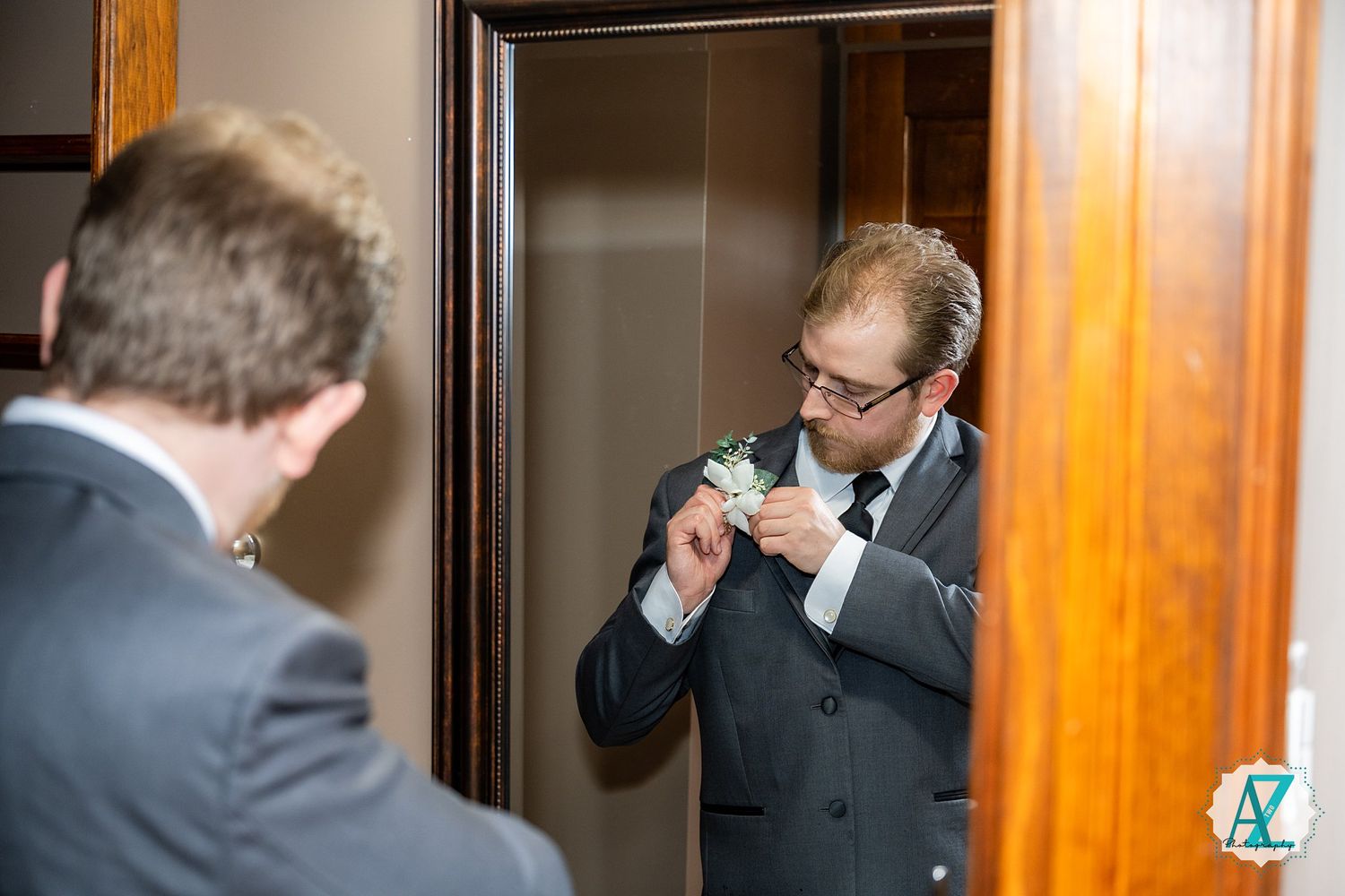 A businessman adjusts his tie while looking in a wooden-framed mirror in a formal setting.