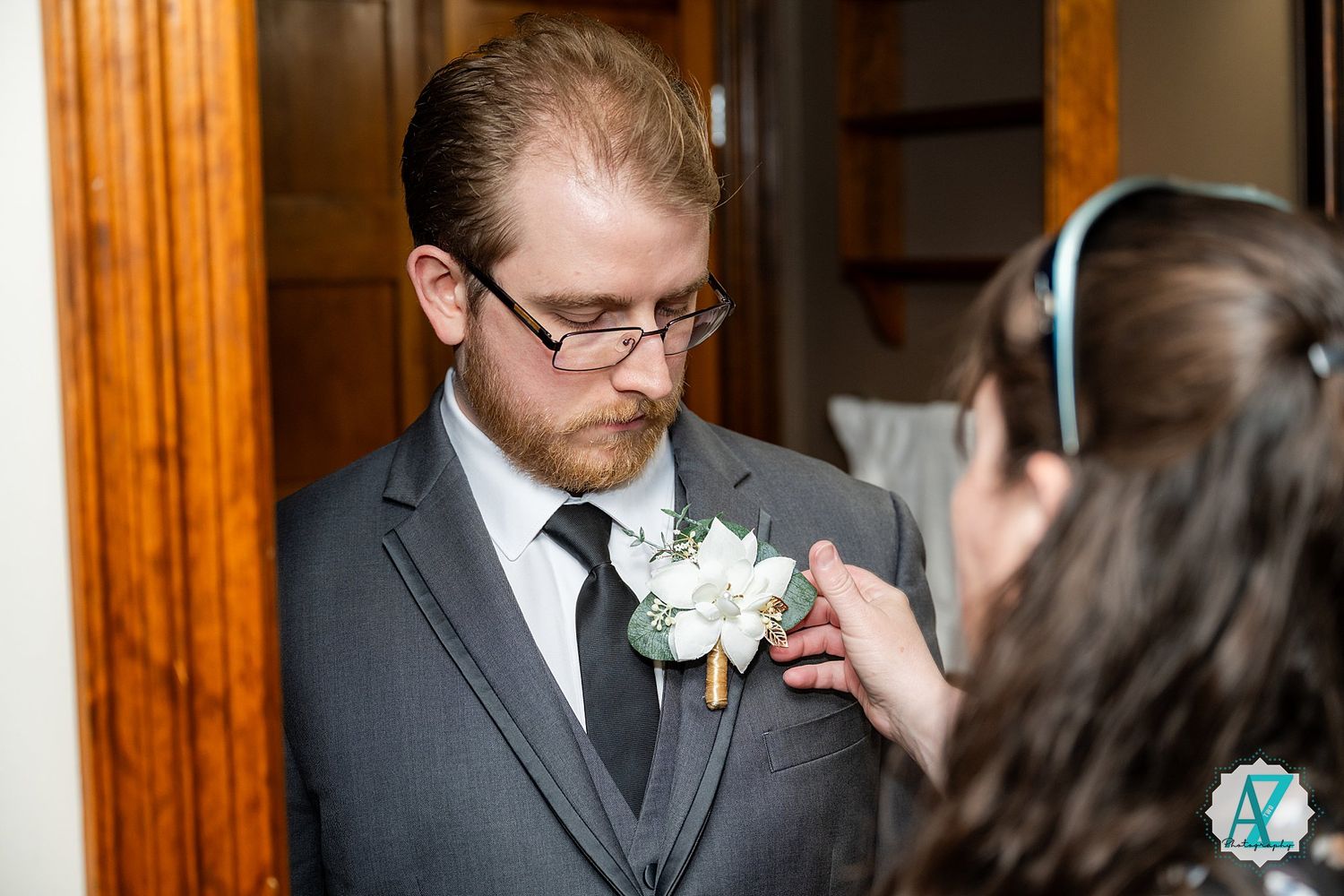 A groom in grey suit getting a white boutonniere flower pinned to his lapel during wedding preparations.