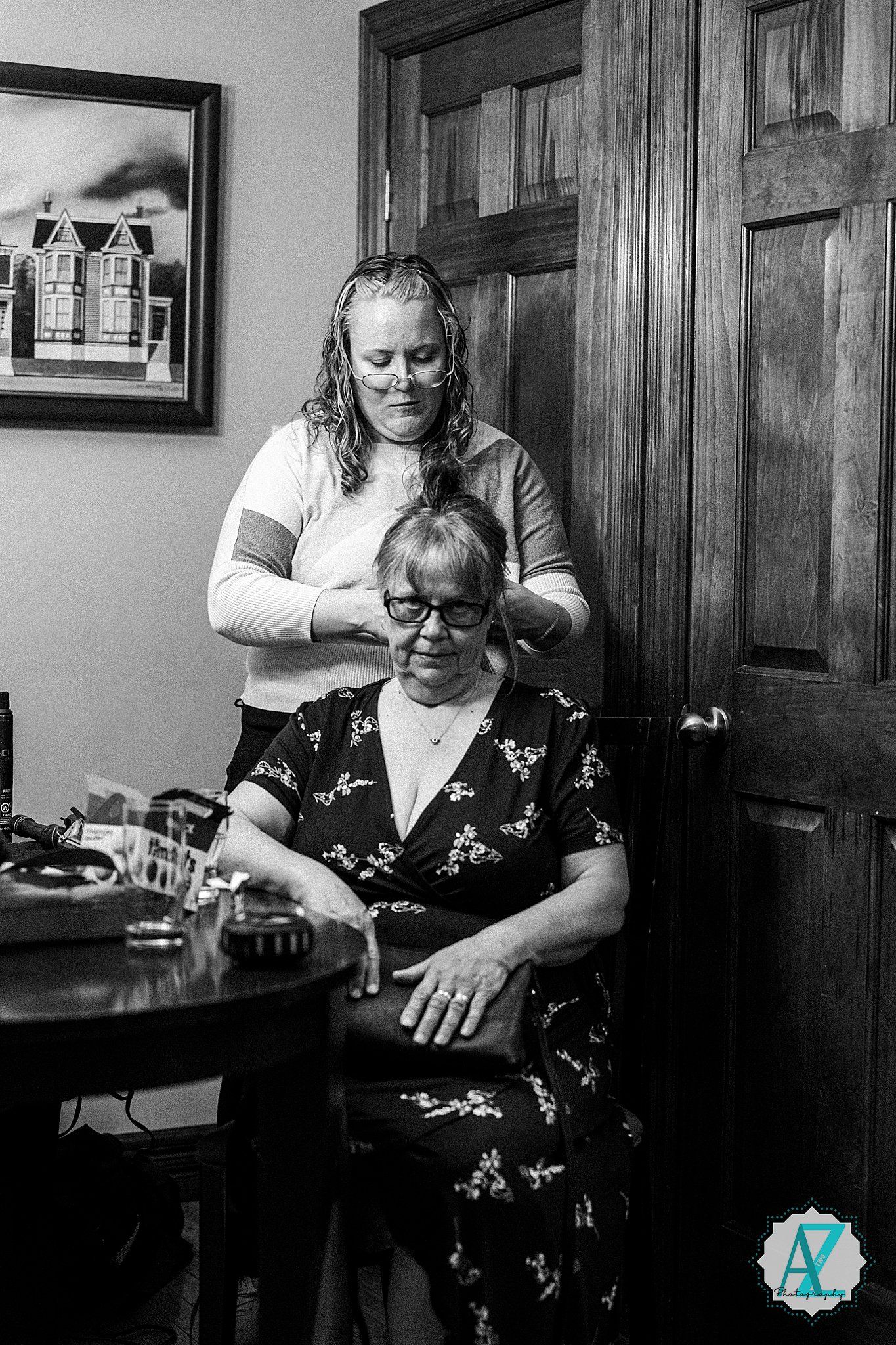 A tender black and white moment shows someone having their hair styled while seated at a wooden table in a cozy interior.