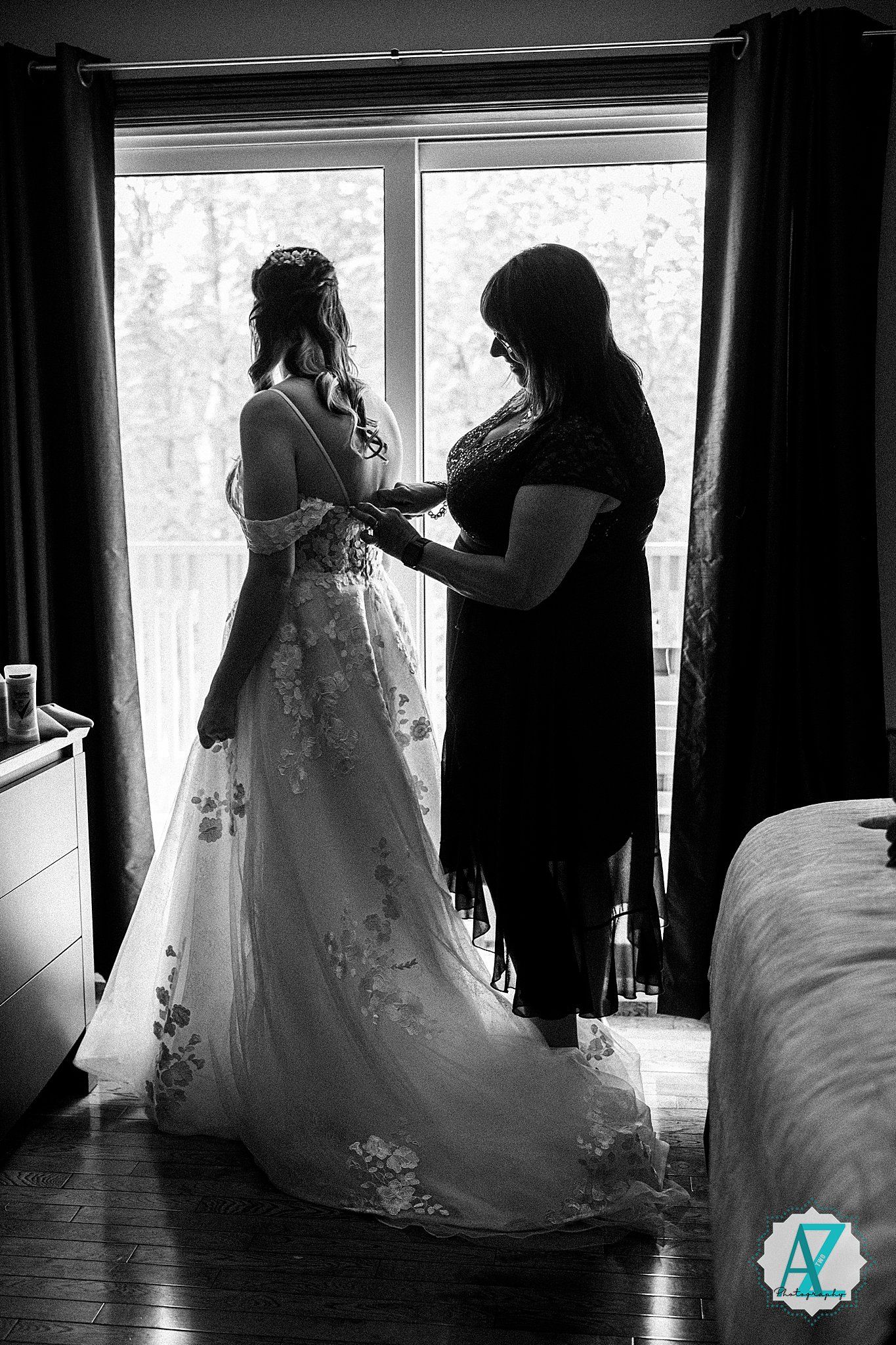 A silhouetted figure helps adjust a wedding dress near a window in black and white photography.