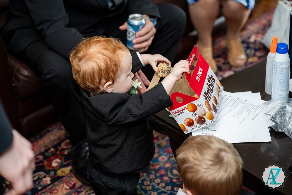 Small children in dark clothing reach for a box of snacks at a gathering with drinks and papers on the table.