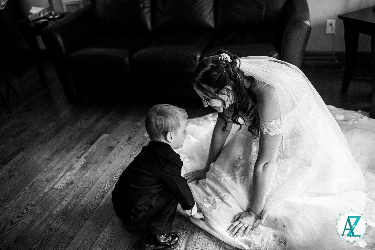 A touching black and white wedding moment as bride in white gown leans down on hardwood floor.
