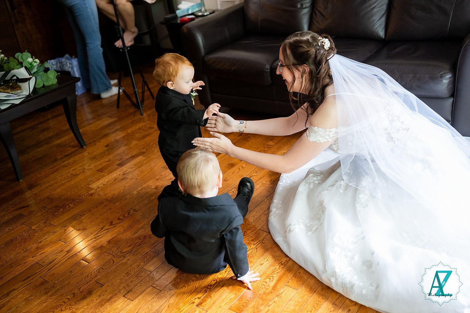 A person in a white wedding dress kneels on hardwood floor interacting with two small children.