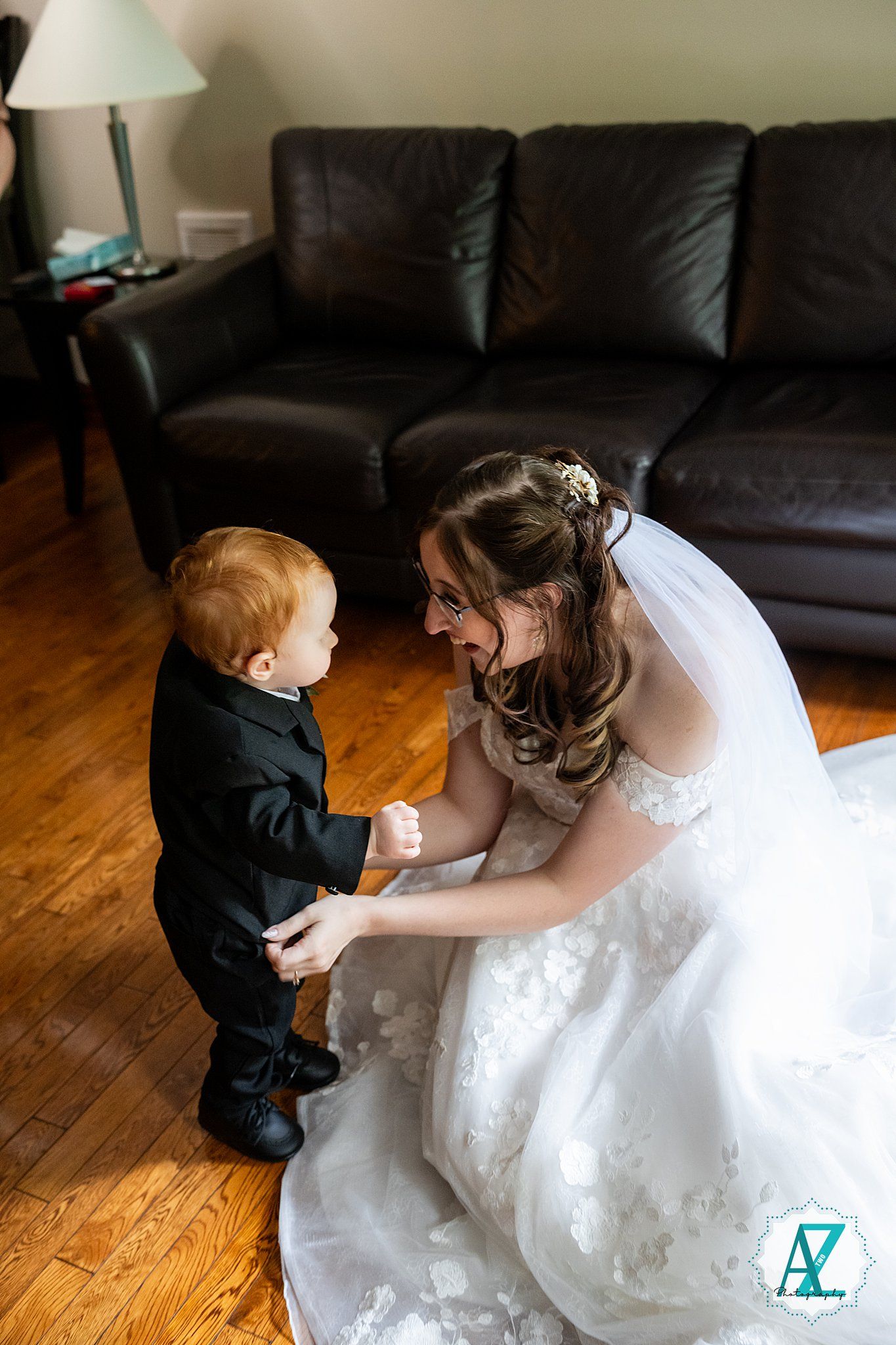 A woman in a white wedding dress kneels on hardwood floor while interacting with a small child in formal attire.