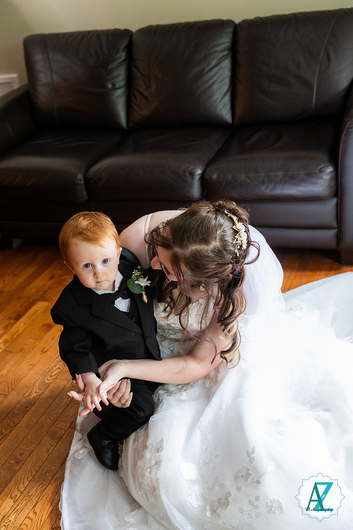 Wedding portrait on hardwood floor with bride in white gown leaning toward small child in formal black attire.