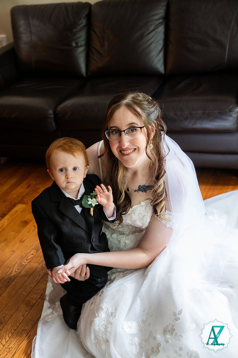 A bride in a white dress and glasses sits on hardwood flooring with a young ring bearer in a black suit.