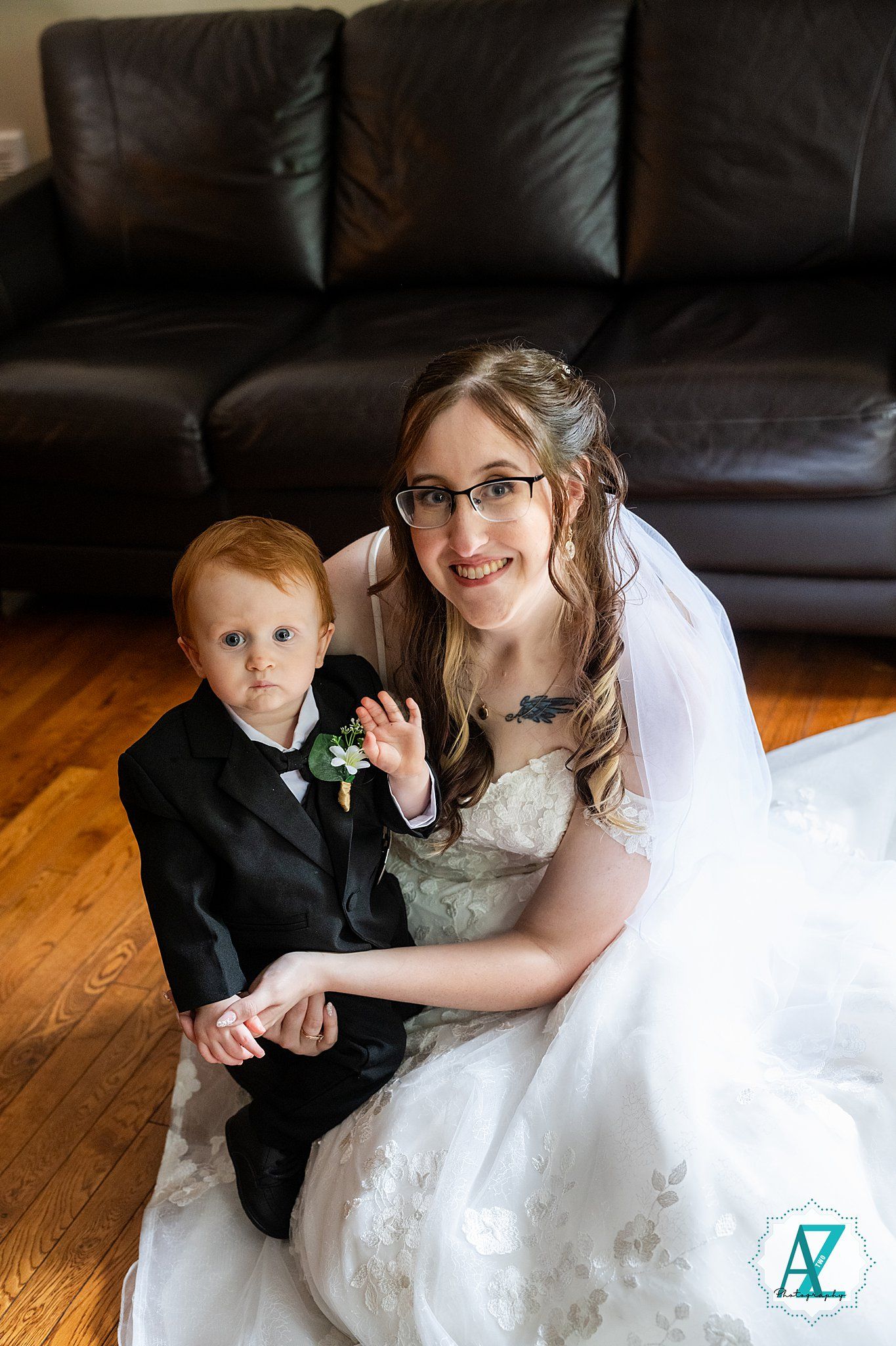 A bride in a white dress and glasses sits on hardwood flooring with a young ring bearer in a black suit.