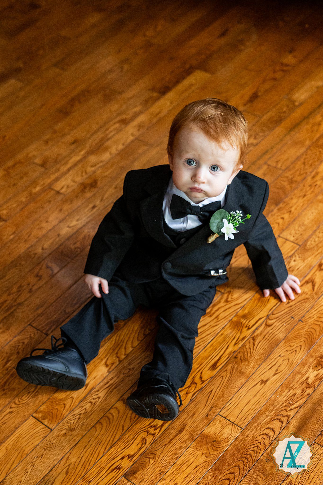 Small child wearing formal black tuxedo lies on wooden floor in series of dynamic poses for wedding photos.