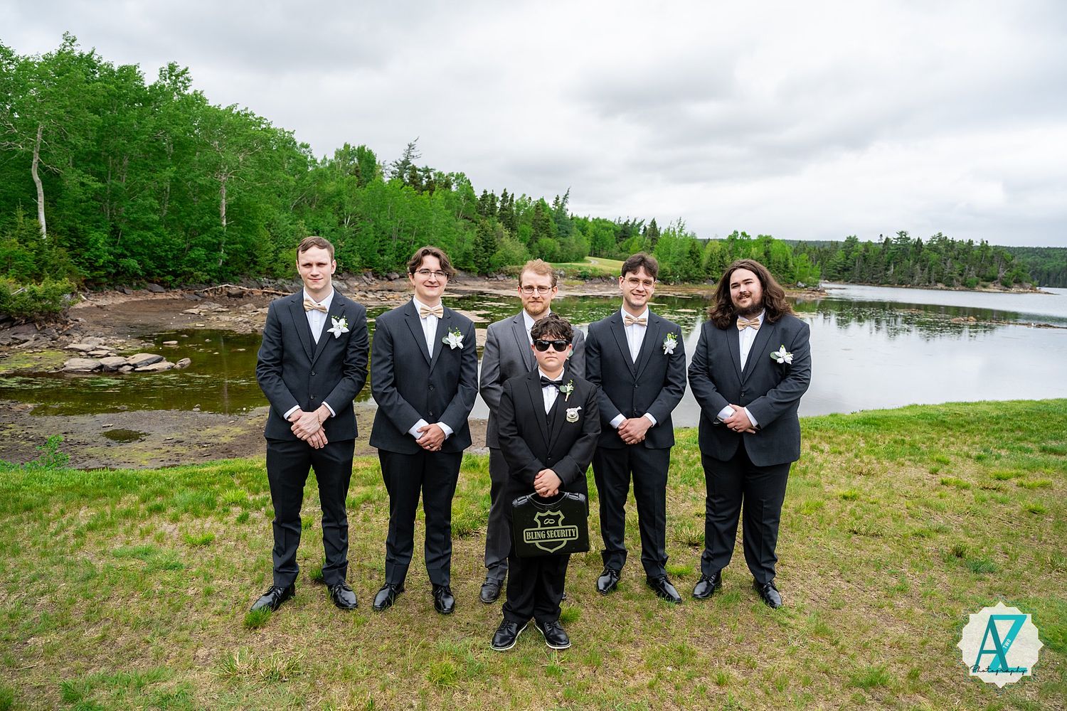 Wedding party in black suits pose for a formal group photo outdoors by a serene lake surrounded by green trees.