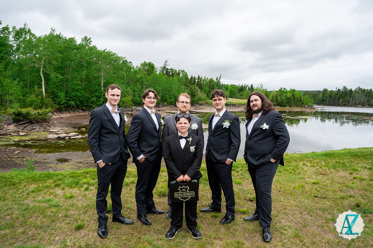 Five people in formal black suits pose together on grassy shore with lake and forest in background.