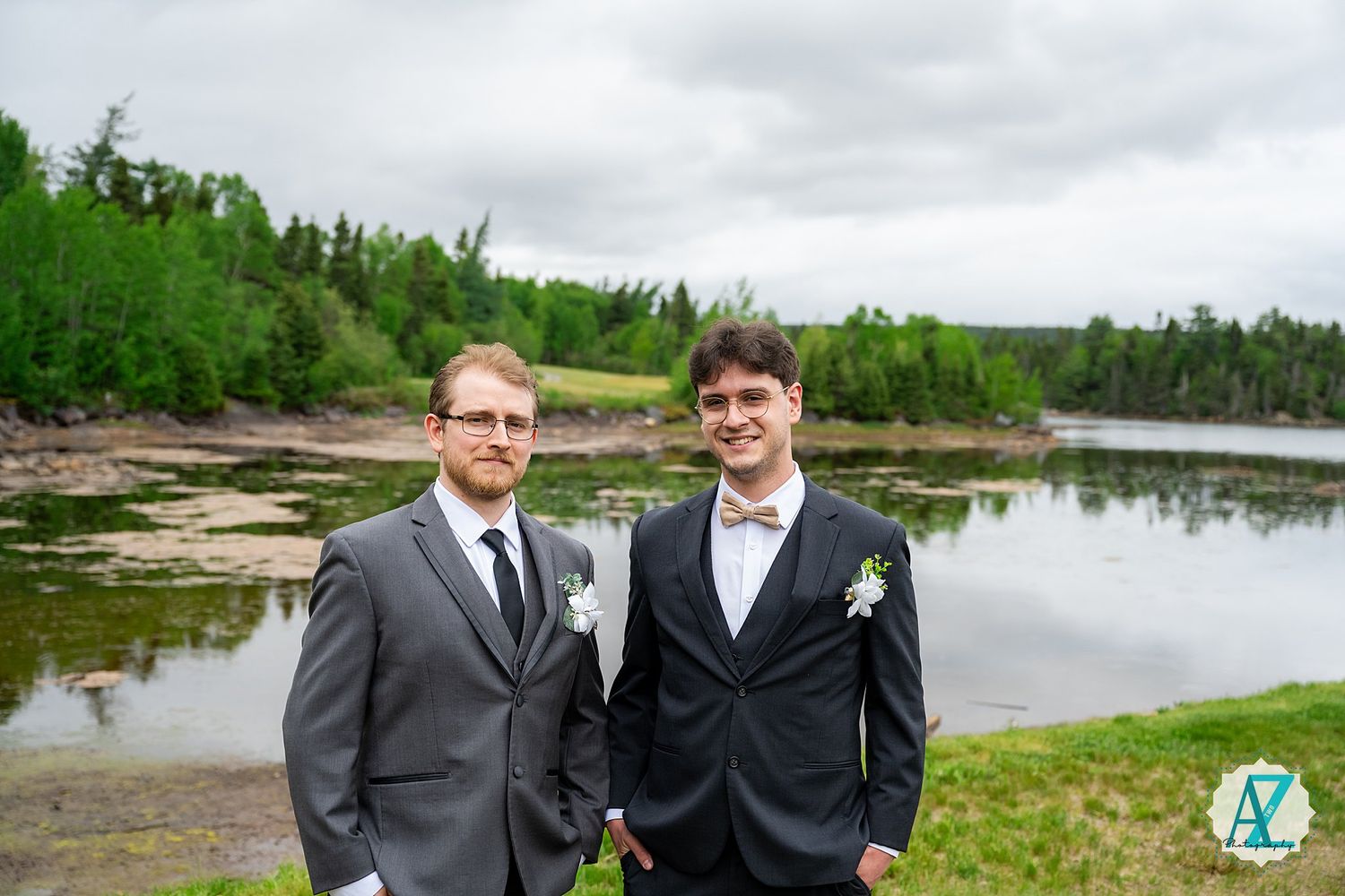 Two people in formal suits stand together on a rocky shoreline with a scenic lake and forest in the background.