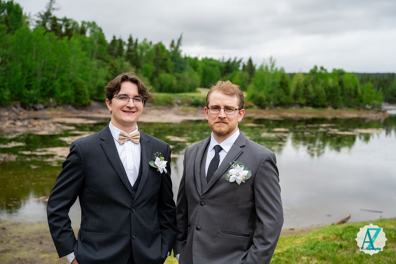 Two dapper looking groomsmen in suits and boutonnieres pose by a scenic waterfront with green forest backdrop.
