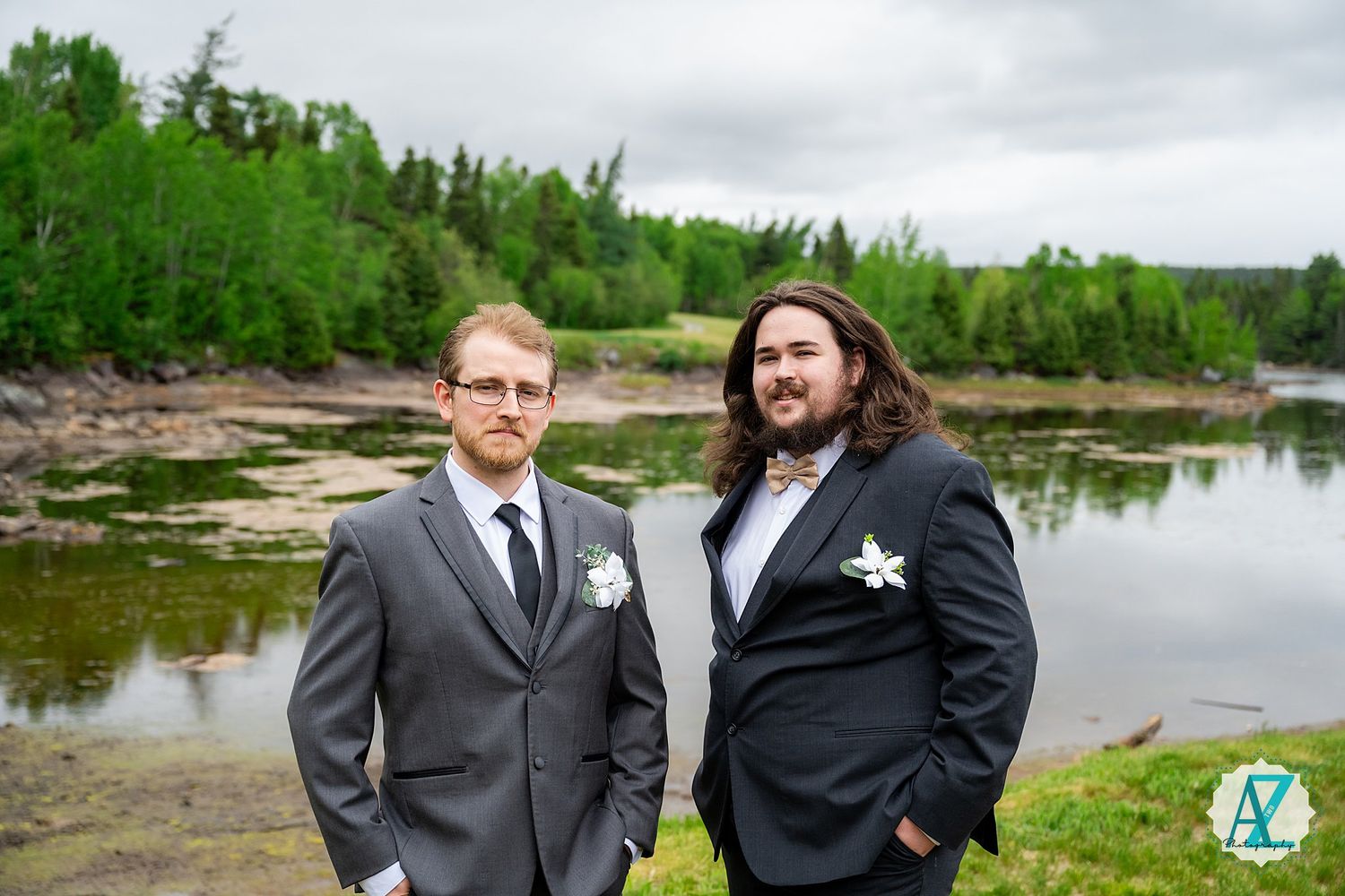 Two people in formal attire stand together by a scenic lake surrounded by evergreen forest on an overcast day.