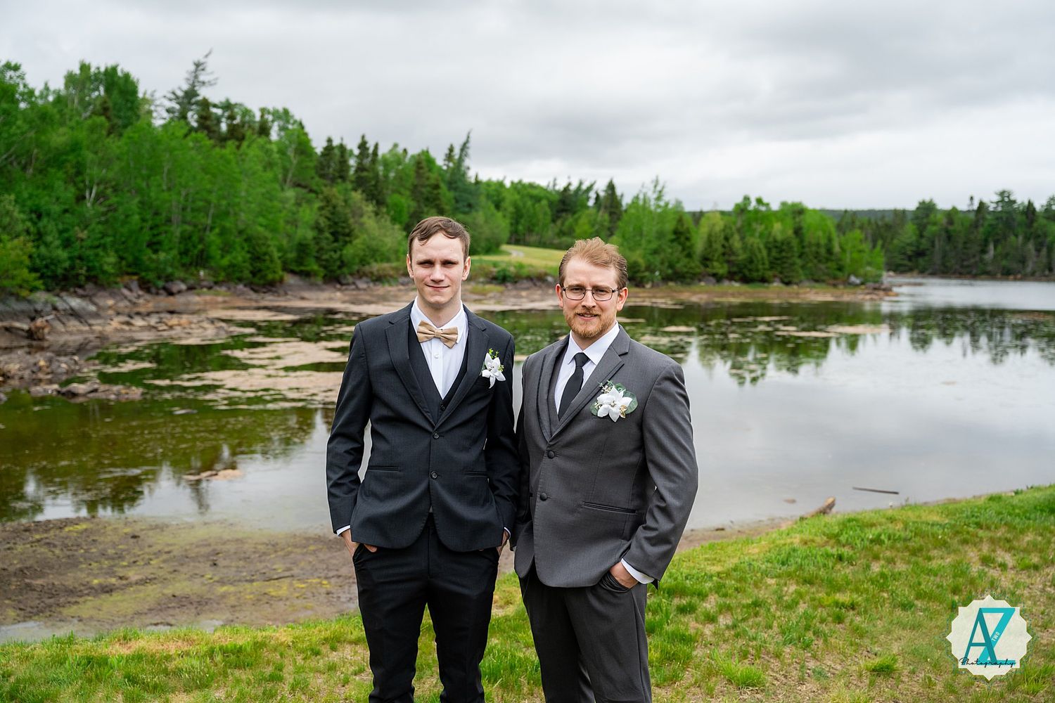 Two people wearing formal black suits stand outdoors by a scenic lake surrounded by green forest.