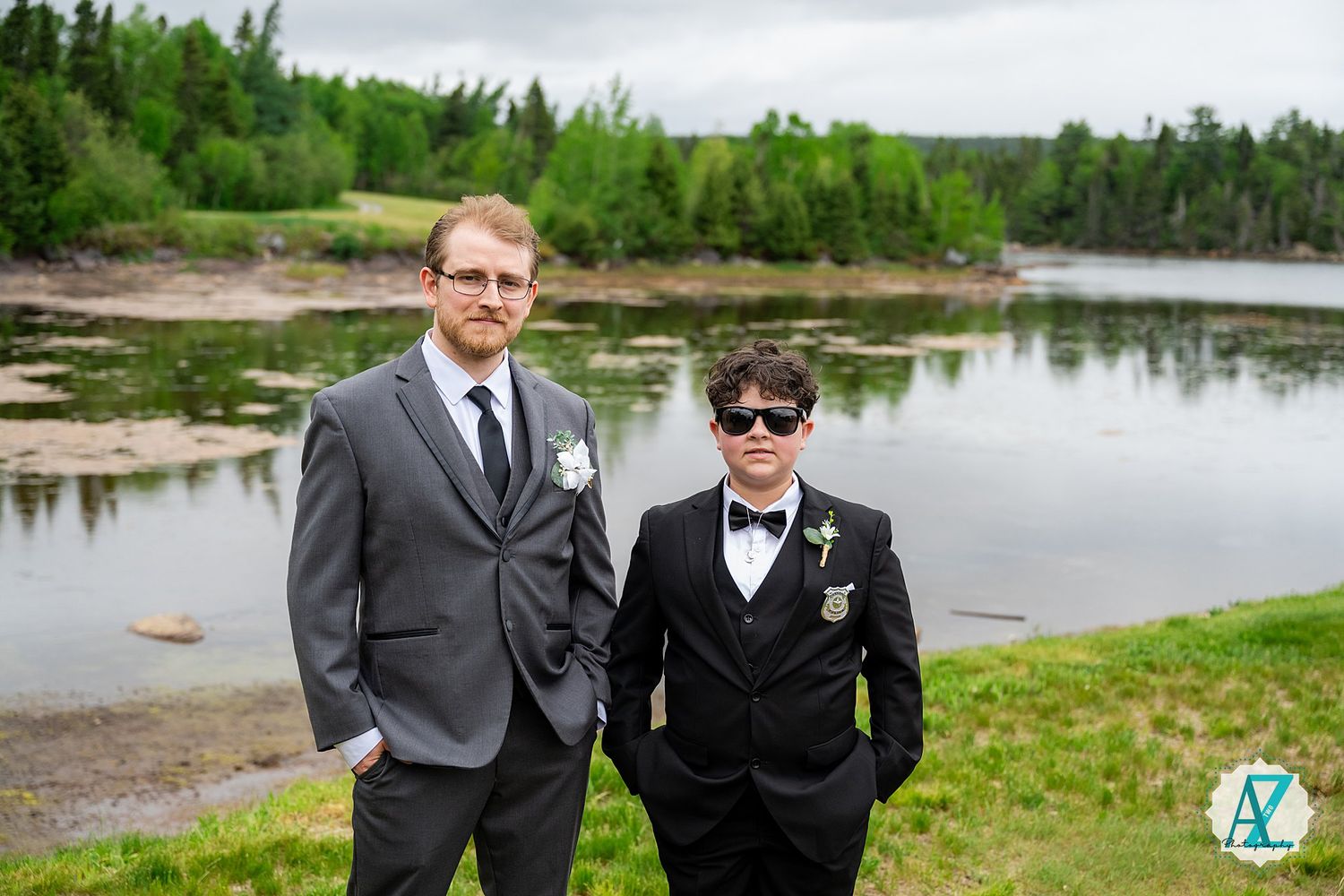 Two people in formal attire stand together by a scenic riverside with green forest and flowing water in the background.
