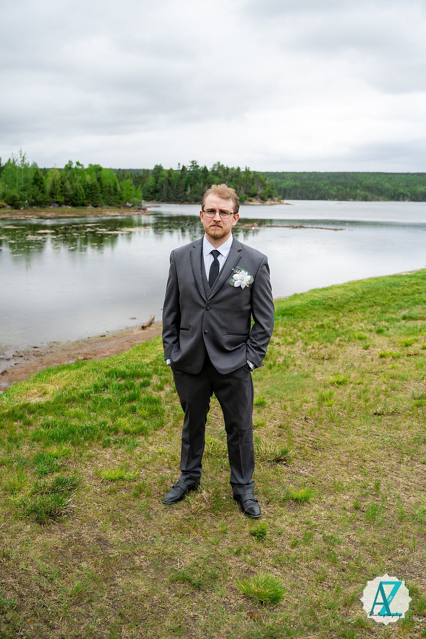 Black-suited figure stands on grassy shoreline overlooking expansive lake under cloudy skies.