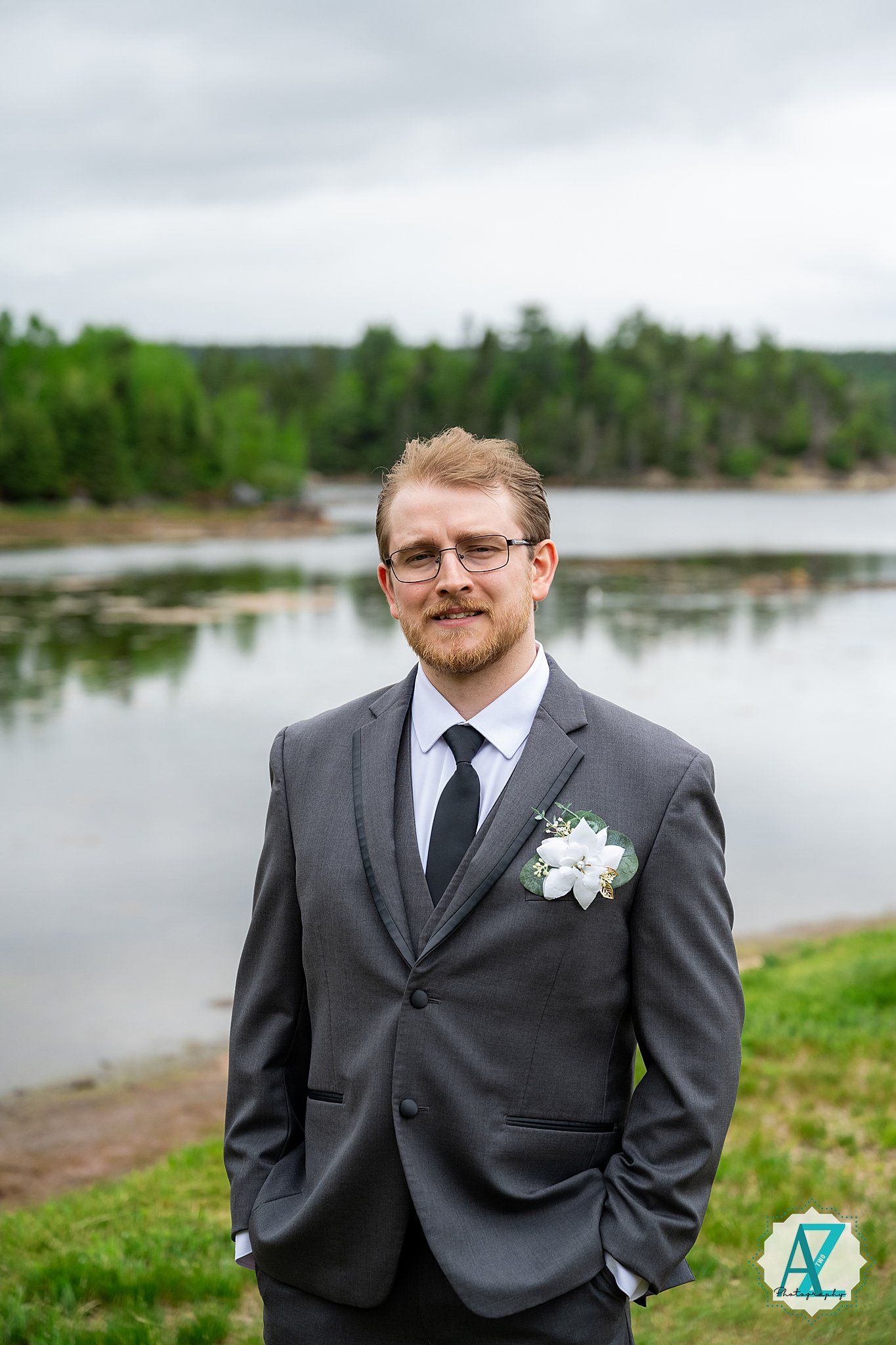 A groom in a dark gray suit and tie with white boutonniere poses outdoors by a scenic lake and forest view.