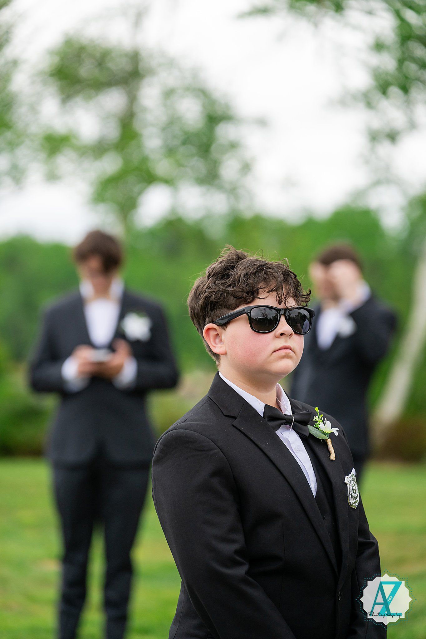 A young person in a black tuxedo and sunglasses strikes a cool pose outdoors during a formal event.