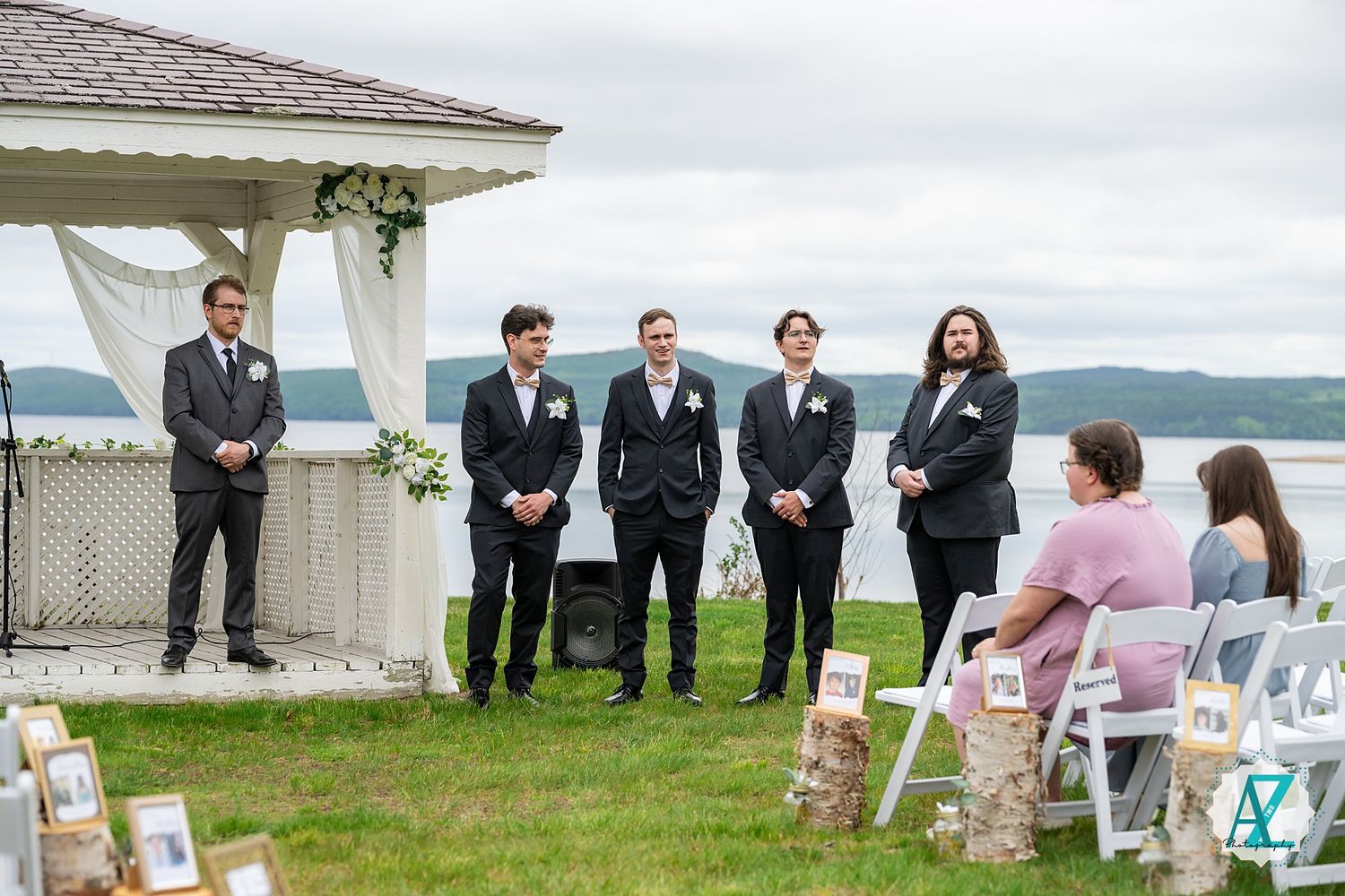Wedding party members stand on grass near white chairs during an outdoor lakeside ceremony setup.