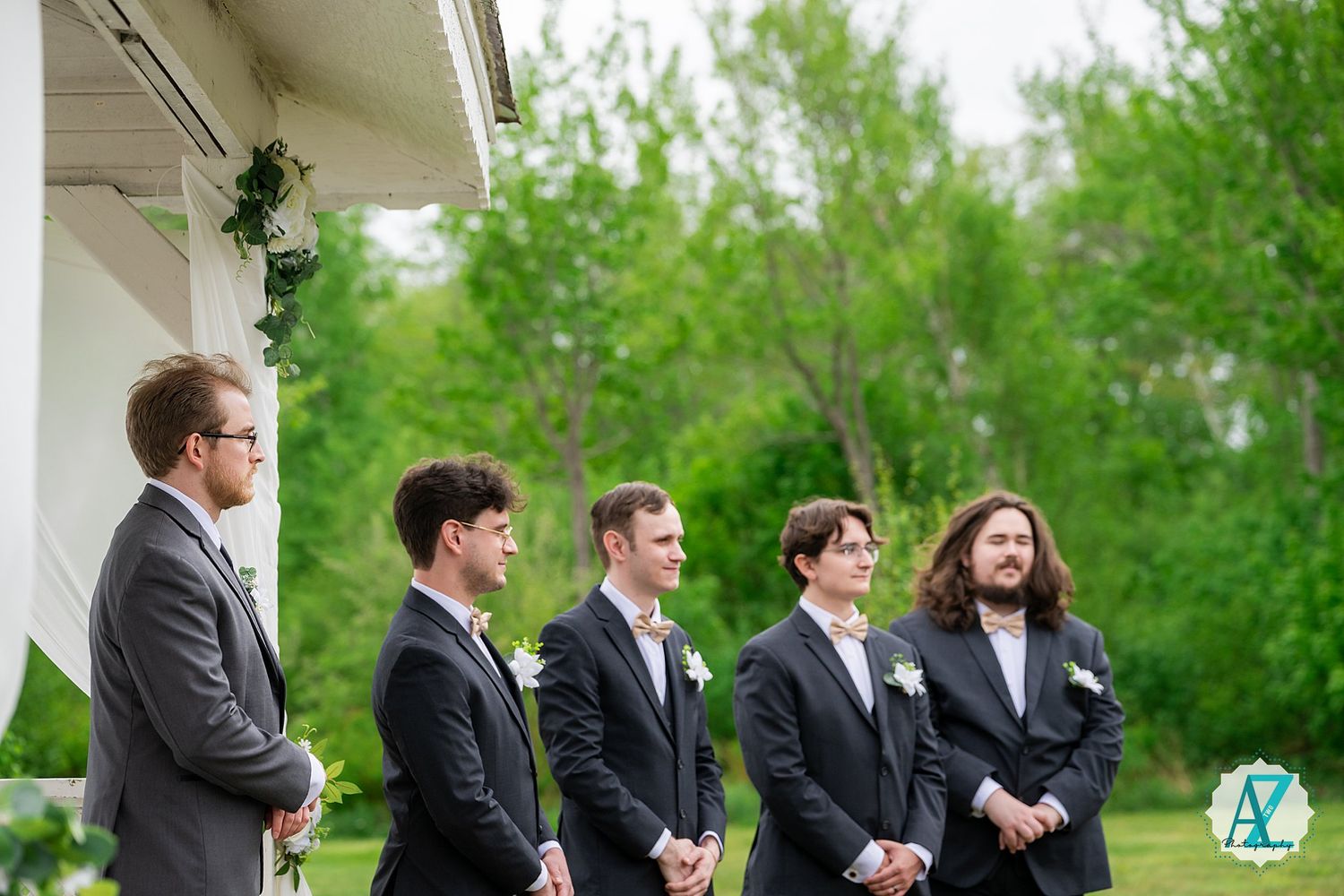 A line of groomsmen in black suits stand outdoors during a wedding ceremony with green trees in the background.