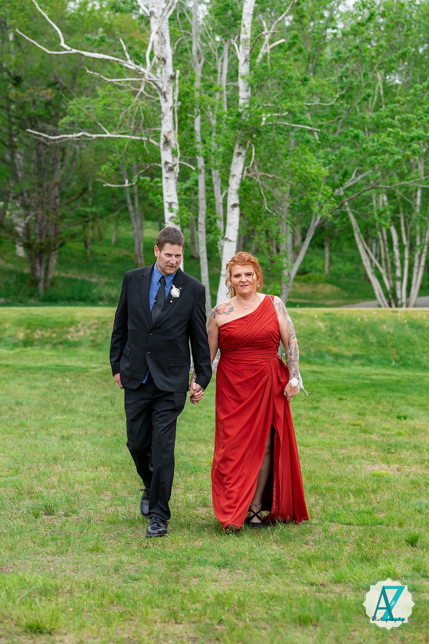 A formal couple walks hand in hand through a grassy area with white birch trees in the background during a special occasion.