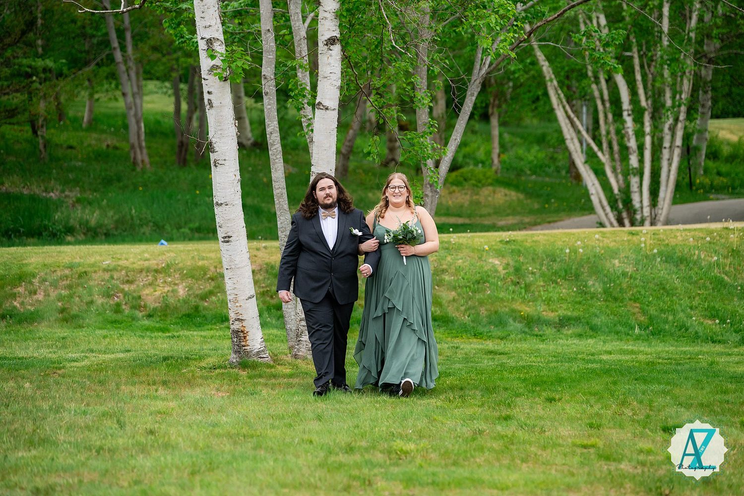 A formal couple in evening attire walks together on a grassy lawn with white birch trees in the background.