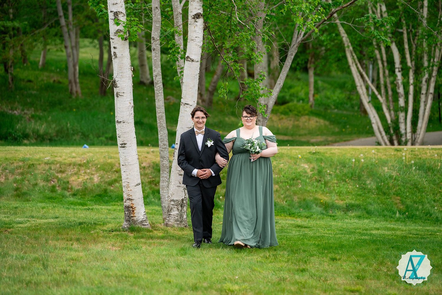 A couple walks arm in arm on lush grass with white birch trees creating a scenic forest backdrop at an outdoor event.