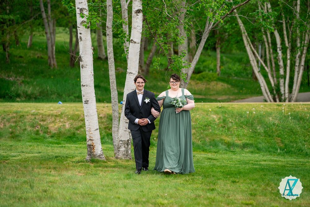 A couple walks arm in arm on lush grass with white birch trees creating a scenic forest backdrop at an outdoor event.