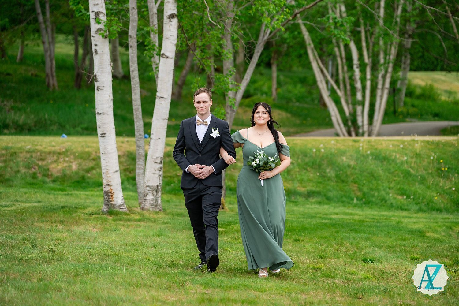 Couple in formal attire walking together between white birch trees on a lush green lawn.