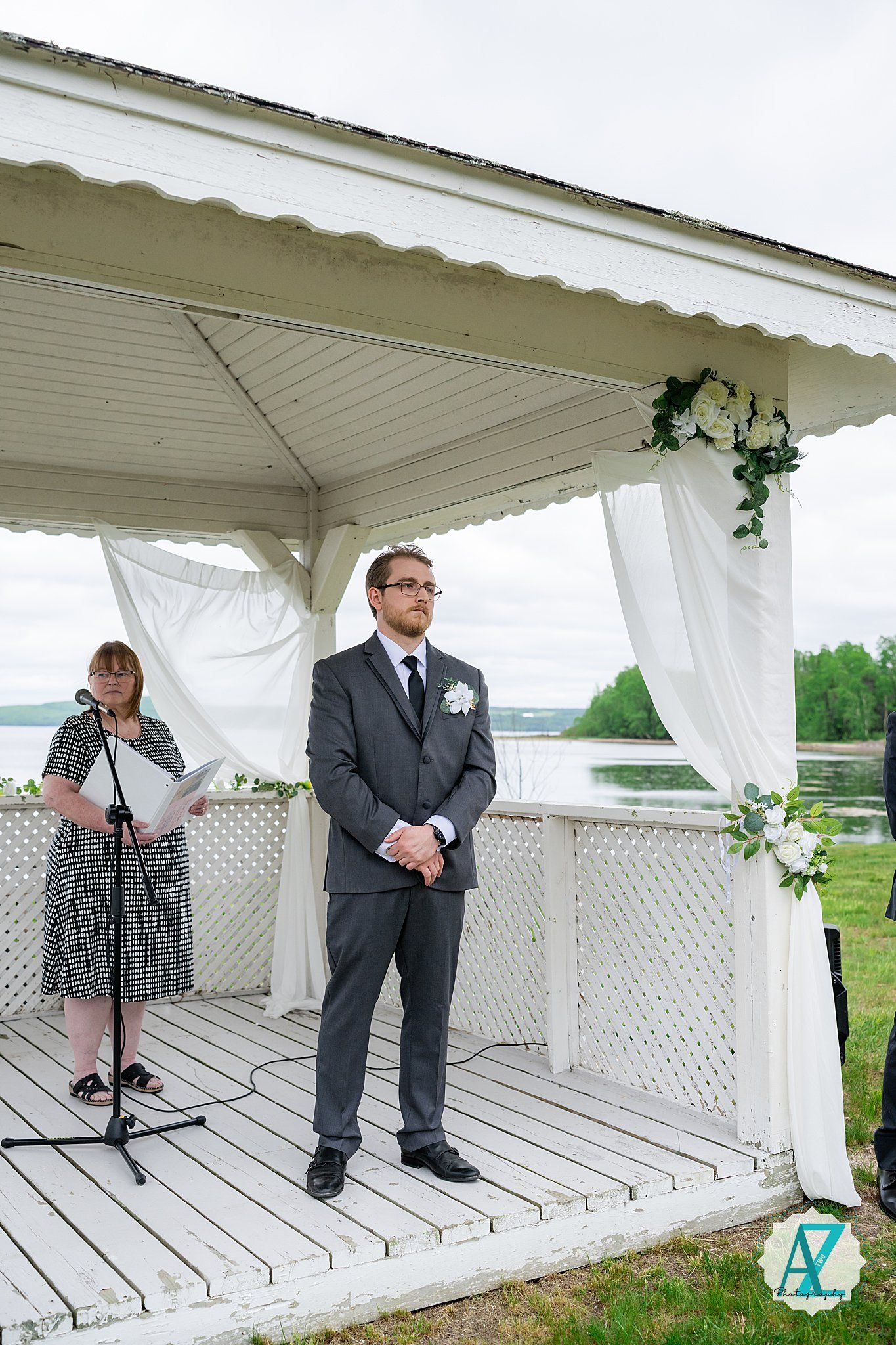 A formal wedding ceremony takes place in a white wooden gazebo overlooking a waterfront setting.