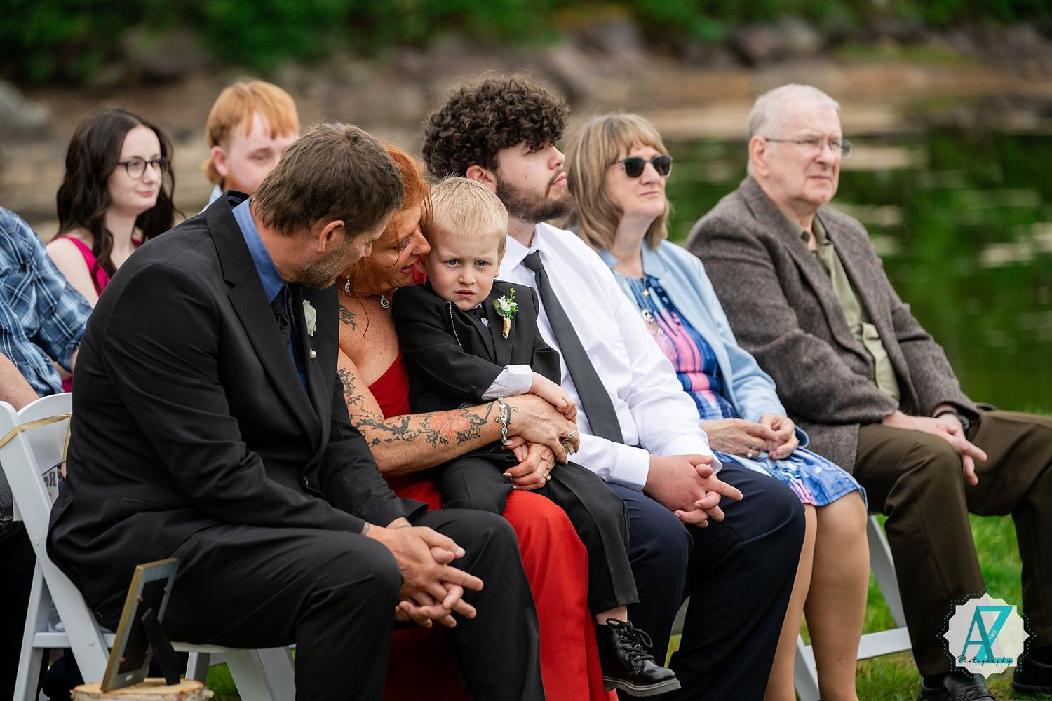 Wedding guests are seated outdoors at a ceremony with lush greenery in the background.