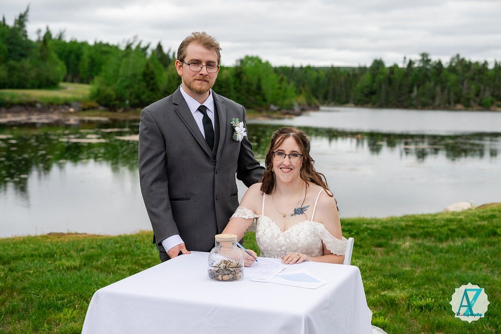 A couple poses behind a white signing table at their lakeside wedding ceremony with forest scenery in the background.