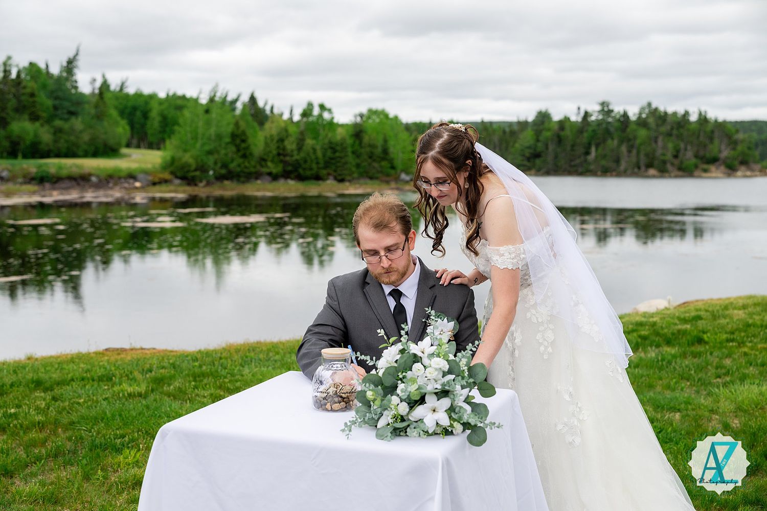 A wedding couple poses by a serene lake surrounded by green trees and grass during an outdoor ceremony.
