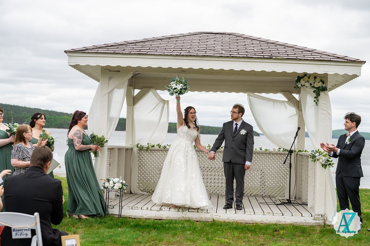Wedding couple celebrates with guests under a decorated white gazebo surrounded by flowers on a cloudy day.